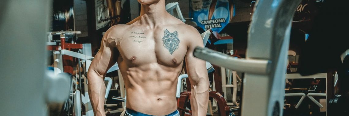 Shirtless man with tattoos posing in a gym environment, showcasing fitness and strength.